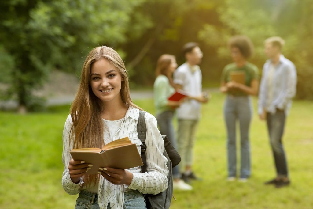 Besutiful college student girl with book posing outdoors with classmates on background